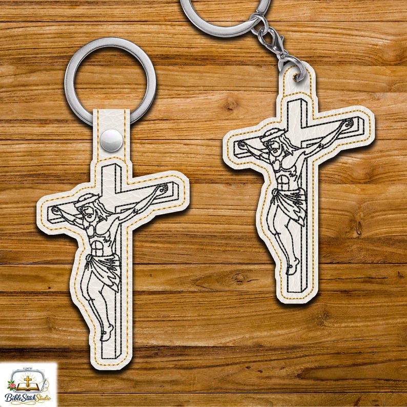 May include: Two keychains featuring a cross with a depiction of Jesus Christ. The cross and figure are outlined in black stitching on a white background, with a gold-colored border. Each keychain has a silver-colored ring.