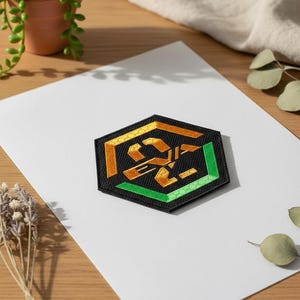 Puede incluir: Un parche hexagonal negro con detalles dorados y verdes. El parche presenta un diseño dorado, con patrones geométricos verdes y dorados. El parche está sobre una superficie blanca.