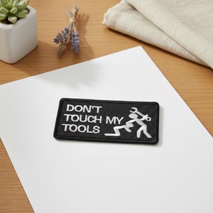 Puede incluir: Parche rectangular negro con texto bordado blanco « DON'T TOUCH MY TOOLS ». El parche presenta un gráfico de dos figuras de palo, una de ellas con una llave inglesa. El parche está sobre una superficie blanca.