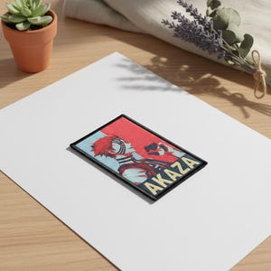 Könnte beinhalten: Gestickter Aufnäher mit einer Charakterillustration. Der Aufnäher hat einen roten und blauen Hintergrund mit dem Namen "AKAZA" in gelben Buchstaben. Die Figur hat rotes Haar und ein unverwechselbares Gesichtsdesign. Der Aufnäher ist rechteckig mit einem schwarzen Rand.