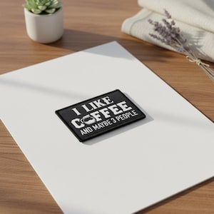 Op de afbeelding: Een zwart rechthoekig embleem met witte tekst die luidt "I LIKE COFFEE AND MAYBE 3 PEOPLE". Het embleem heeft een koffiepotafbeelding. Het embleem ligt op een wit oppervlak.