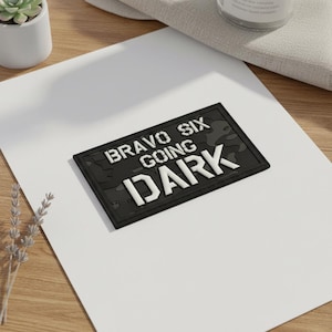 Könnte beinhalten: Ein schwarzer rechteckiger Aufnäher mit dem Text "BRAVO SIX GOING DARK" in weißen, fetten Großbuchstaben. Der Hintergrund hat ein Tarnmuster. Der Aufnäher liegt auf einer weißen Oberfläche.