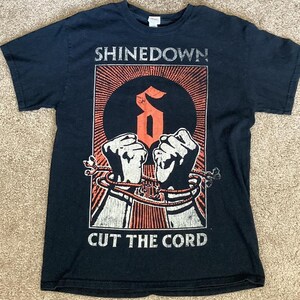 May include: A navy blue t-shirt featuring the band name "SHINEDOWN" above a graphic of two clenched fists bound by rope. The text "CUT THE CORD" is printed below the graphic. The design is in shades of orange, white, and black.