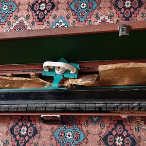 Op de afbeelding: Een vintage breimachine met een zwarte naaldenbalk en een talingkleurige slede, gehuisvest in een bruine, getextureerde draagkoffer met een groene binnenvoering. De set bevat bruin beschermpapier voor handmatige textielproductie.