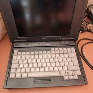 Peut inclure: Un ordinateur portable vintage Canon BN700 doté d'un châssis gris foncé et d'un clavier QWERTY gris clair. L'appareil comprend un écran noir, un bouton trackpoint central sur le clavier et une rangée de touches de fonction en haut.