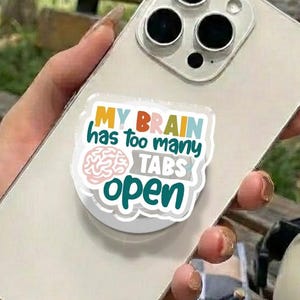 May include: A silver smartphone with a white pop-up grip featuring the text "MY BRAIN has too many TABS open" in a colorful design. The phone is held in a hand, with a bracelet visible. Other accessories are in the background.