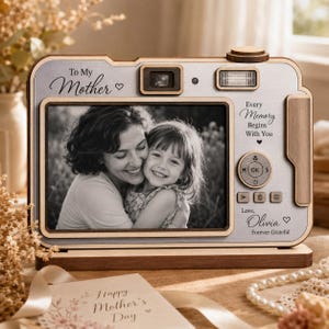 May include: A wooden camera-shaped photo frame with a light-colored faceplate featuring the text To My Mother, Every Memory Begins With You, and Love, Olivia. The frame displays a black and white photograph of an adult and a child embracing.
