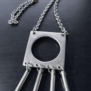 May include: A silver-toned necklace with a square pendant featuring a circular cutout. The pendant has four cylindrical charms hanging from the bottom. The necklace chain is a classic link style.