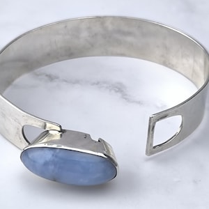 May include: A silver-toned cuff bracelet with a light blue oval gemstone. The bracelet has a wide band with a geometric cutout on one end and a clasp with the gemstone on the other. The gemstone is approximately 1 inch long.