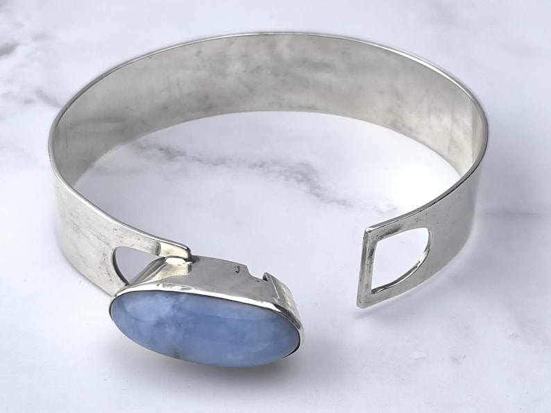 May include: A silver-toned cuff bracelet with a light blue oval gemstone. The bracelet has a wide band with a geometric cutout on one end and a clasp with the gemstone on the other. The gemstone is approximately 1 inch long.