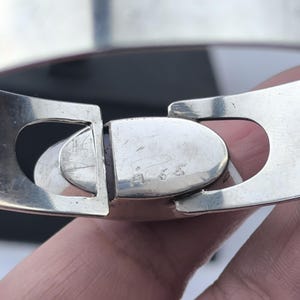 May include: Close-up of a silver-toned hinged bracelet clasp. The clasp is oval-shaped and connects two wide, flat bands. The metal has a polished finish with some visible wear. The bracelet appears to be a piece of jewelry.
