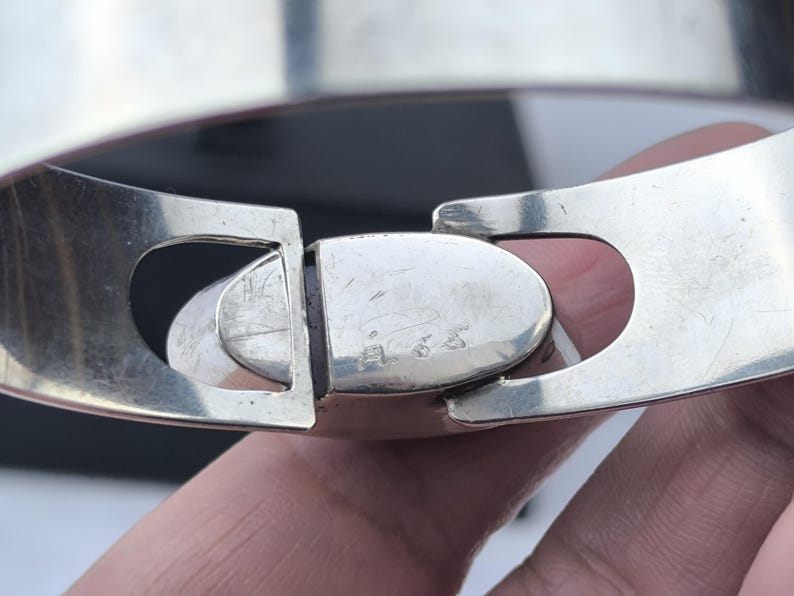 May include: Close-up of a silver-toned hinged bracelet clasp. The clasp is oval-shaped and connects two wide, flat bands. The metal has a polished finish with some visible wear. The bracelet appears to be a piece of jewelry.