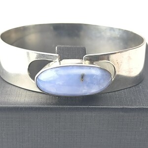 May include: A silver cuff bracelet featuring a large, oval, pale blue gemstone. The bracelet has a wide, flat band with cut-out details on each side of the gemstone. The gemstone is set in a silver bezel.