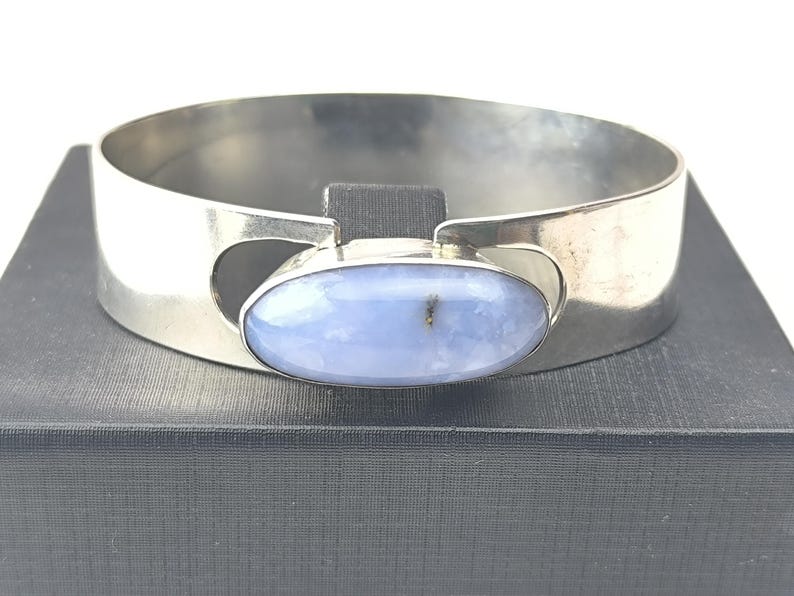 May include: A silver cuff bracelet featuring a large, oval, pale blue gemstone. The bracelet has a wide, flat band with cut-out details on each side of the gemstone. The gemstone is set in a silver bezel.