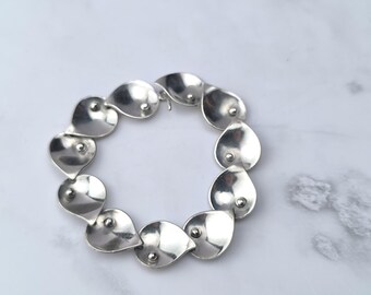 May include: A silver-toned bracelet featuring a series of teardrop-shaped links. Each link has a small, raised detail at its center. The bracelet is connected with a simple clasp, and is displayed on a white surface.