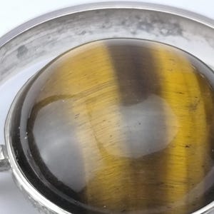 Peut inclure: Gros plan d'un pendentif argent&eacute; avec une grosse pierre &oelig;il de tigre ronde. La pierre est de couleur brun dor&eacute; chaud avec des stries naturelles. Le pendentif a un design circulaire.