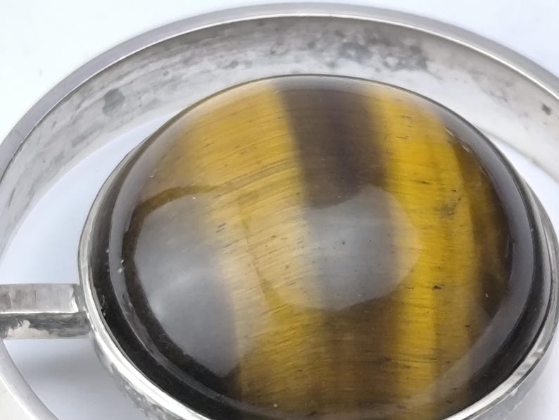 Peut inclure: Gros plan d'un pendentif argent&eacute; avec une grosse pierre &oelig;il de tigre ronde. La pierre est de couleur brun dor&eacute; chaud avec des stries naturelles. Le pendentif a un design circulaire.