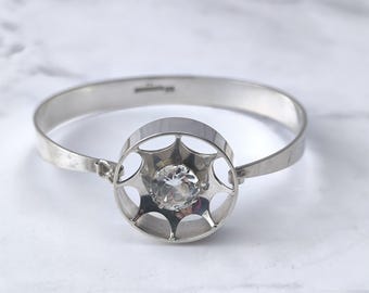 May include: A silver-toned bangle bracelet with a central circular design. The circle features an openwork pattern surrounding a clear, faceted gemstone. The bracelet has a simple, elegant design, suitable for everyday wear or special occasions.