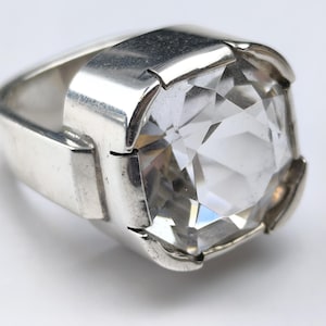 May include: A silver-toned ring featuring a large, round, faceted clear stone set within a thick, polished metal band. The setting holds the stone securely with four prominent prongs, showcasing a reflective surface and a geometric design.