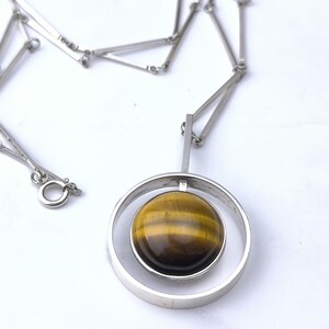 Peut inclure: Collier argent&eacute; avec un pendentif circulaire contenant une pierre &oelig;il de tigre polie et ronde. La pierre pr&eacute;sente des bandes marron et dor&eacute;es. La cha&icirc;ne est compos&eacute;e de maillons rectangulaires et poss&egrave;de un fermoir. Le pendentif mesure environ 3 cm de diam&egrave;tre.