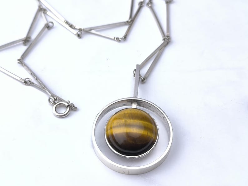 Peut inclure: Collier argent&eacute; avec un pendentif circulaire contenant une pierre &oelig;il de tigre polie et ronde. La pierre pr&eacute;sente des bandes marron et dor&eacute;es. La cha&icirc;ne est compos&eacute;e de maillons rectangulaires et poss&egrave;de un fermoir. Le pendentif mesure environ 3 cm de diam&egrave;tre.