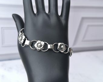 May include: A silver bracelet featuring a floral design. The bracelet has multiple flower-shaped charms connected by circular links and rectangular bars. The flowers have detailed petals and a central bead. The bracelet is displayed on a black mannequin hand.