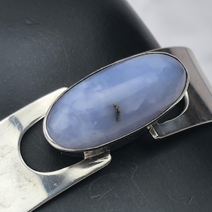 May include: A silver bracelet featuring a large, oval, pale blue gemstone. The gemstone is set in a silver bezel and is the focal point of the bracelet. The bracelet has a simple, modern design.