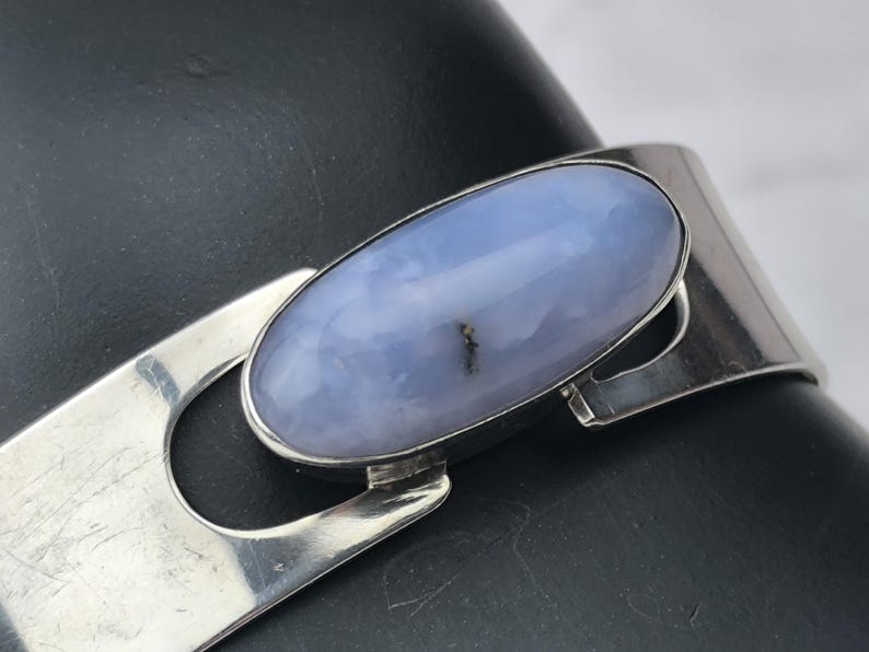 May include: A silver bracelet featuring a large, oval, pale blue gemstone. The gemstone is set in a silver bezel and is the focal point of the bracelet. The bracelet has a simple, modern design.