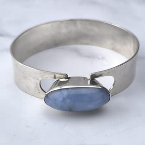 May include: A silver-toned cuff bracelet with a large, oval, pale blue gemstone at the center. The bracelet has a wide band with cut-out details on either side of the gemstone. The gemstone is set in a silver bezel.