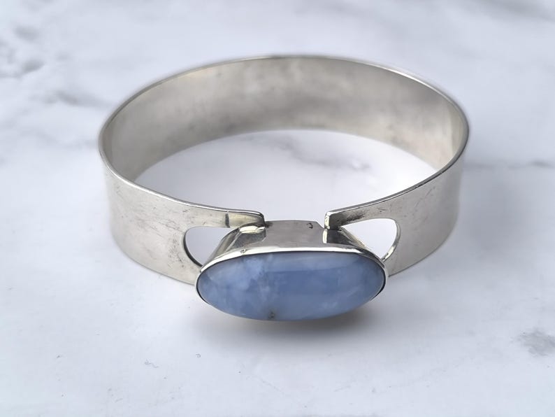 May include: A silver-toned cuff bracelet with a large, oval, pale blue gemstone at the center. The bracelet has a wide band with cut-out details on either side of the gemstone. The gemstone is set in a silver bezel.