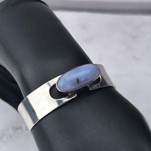 May include: A silver cuff bracelet featuring a light blue oval gemstone. The bracelet has a simple, modern design with a polished finish. The gemstone is set in a bezel setting and has a subtle, natural pattern.
