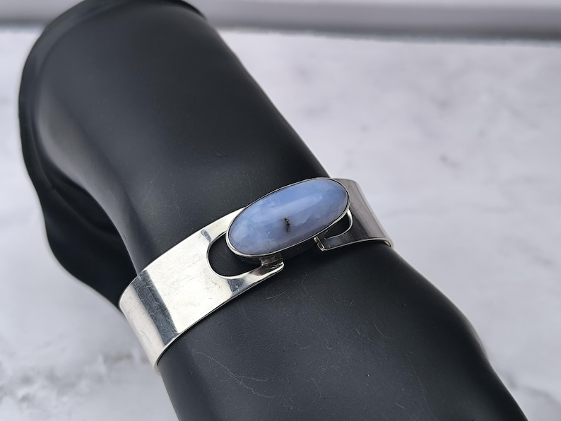 May include: A silver cuff bracelet featuring a light blue oval gemstone. The bracelet has a simple, modern design with a polished finish. The gemstone is set in a bezel setting and has a subtle, natural pattern.