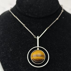 Peut inclure: Un collier en argent avec un pendentif en &oelig;il de tigre. Le pendentif est une pierre polie et ronde, sertie dans un double anneau en argent. La cha&icirc;ne est d&eacute;licate. L'&oelig;il de tigre affiche des tons bruns et dor&eacute;s.