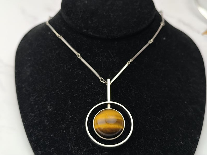 Peut inclure: Un collier en argent avec un pendentif en &oelig;il de tigre. Le pendentif est une pierre polie et ronde, sertie dans un double anneau en argent. La cha&icirc;ne est d&eacute;licate. L'&oelig;il de tigre affiche des tons bruns et dor&eacute;s.