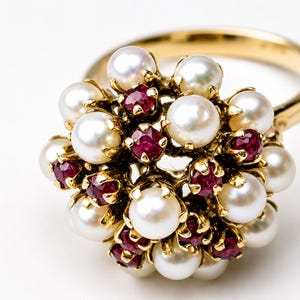 Peut inclure: Une bague en or ornée d'un groupe de perles blanches et de petites pierres précieuses rouges et rondes. Le design de la bague est floral, avec les perles et les pierres précieuses disposées pour ressembler à une fleur.
