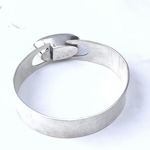 May include: A silver-toned metal bangle bracelet with a rectangular, blue-grey stone. The bracelet has a simple, modern design with a clasp closure. The band measures approximately 2 inches wide.