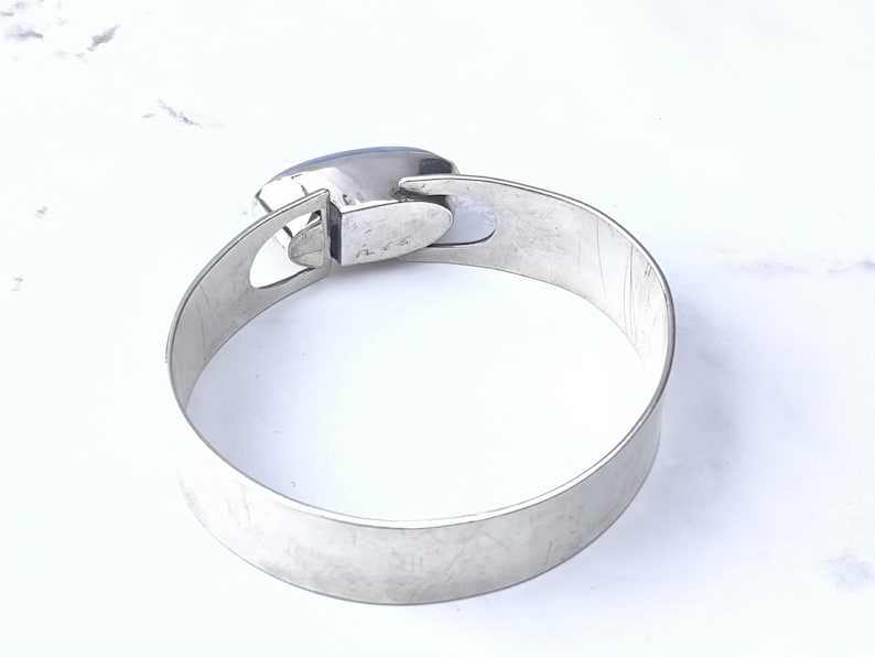 May include: A silver-toned metal bangle bracelet with a rectangular, blue-grey stone. The bracelet has a simple, modern design with a clasp closure. The band measures approximately 2 inches wide.