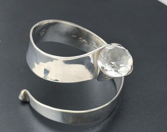 May include: A silver-toned bracelet with a large, clear, faceted gemstone. The bracelet has a spiral design with a wide band that curves around the wrist and tapers to a point. The gemstone is round and set in a bezel.