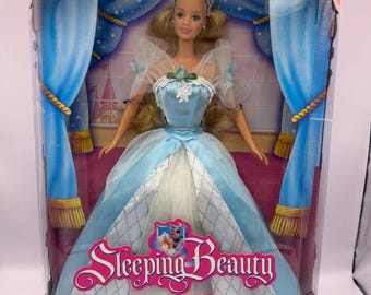 May include: A Sleeping Beauty Barbie doll in a light blue and white gown with a silver tiara, packaged in a box featuring blue curtains and a castle illustration. The box displays the text Sleeping Beauty Barbie and a warning label at the bottom.