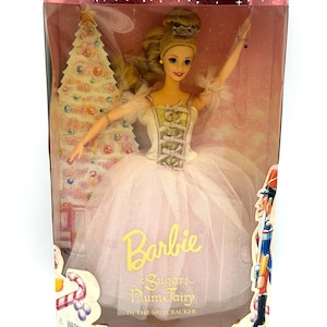 Puede incluir: Una muñeca Barbie Sugar Plum Fairy en una caja de plástico transparente. La muñeca lleva un tutú rosa y un corpiño blanco y dorado. La caja dice "Collector Edition" y "Barbie Sugar Plum Fairy in The Nutcracker."