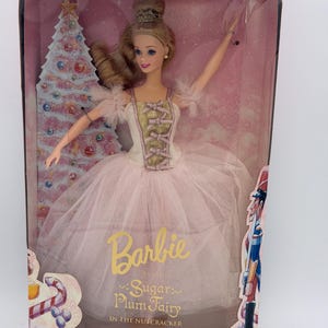 May include: A Barbie doll as the Sugar Plum Fairy in the Nutcracker, packaged in a Collector Edition box. The doll has blonde hair, a gold crown, and wears a cream and gold bodice with a layered pink tulle skirt. The box features a Christmas tree.