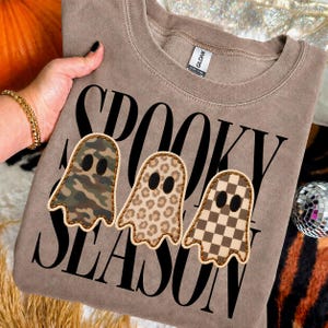 May include: A taupe crewneck sweatshirt featuring the black text SPOOKY SEASON. Three ghost-shaped patches are layered over the text, each with a different pattern: one with green and brown camouflage, one with a tan leopard print, and one with a brown checkerboard.