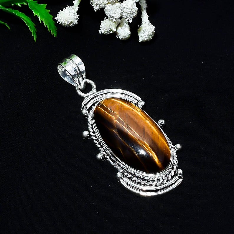 May include: A silver pendant with an oval tiger's eye stone. The stone is a rich brown color with golden streaks. The pendant has a decorative silver border with a loop for a chain. The background is black.