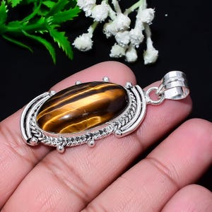 May include: Oval tiger's eye pendant with a silver-colored setting. The gemstone features bands of brown and gold. The pendant has a decorative border and a loop for a chain. Jewelry.