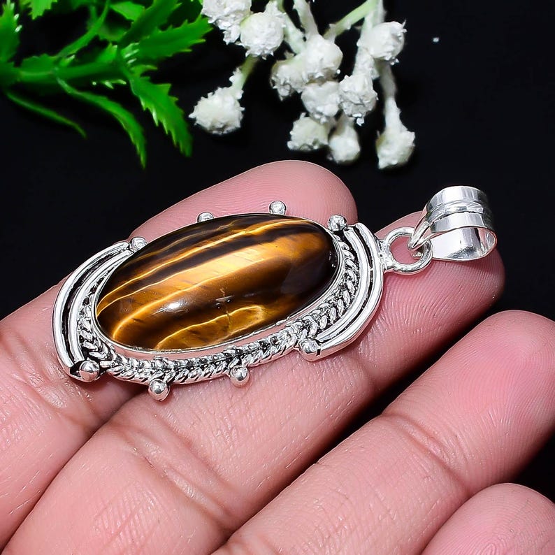 May include: Oval tiger's eye pendant with a silver-colored setting. The gemstone features bands of brown and gold. The pendant has a decorative border and a loop for a chain. Jewelry.