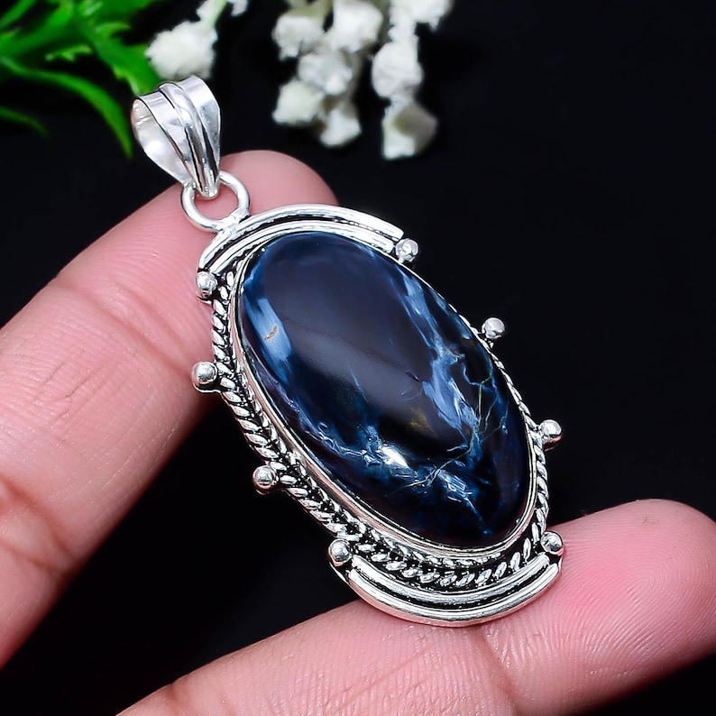 Puede incluir: Colgante ovalado con una piedra preciosa azul oscuro pulida con vetas blancas y doradas. La piedra est&aacute; engastada en un marco de metal plateado con un borde decorativo en forma de cuerda y un lazo para una cadena.