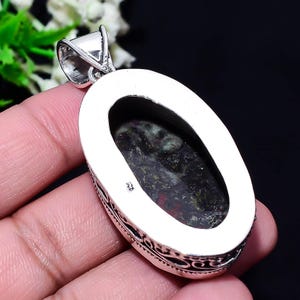 May include: Oval-shaped pendant with a dark, speckled stone set in a silver metal frame. The stone has a mix of dark green, red, and black colors. The silver frame has a decorative pattern around the edge and a loop for a chain.