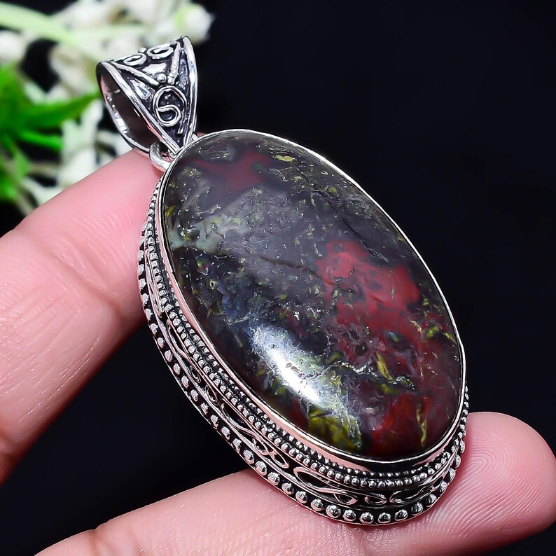 May include: Oval pendant with a large, polished gemstone. The stone features a mix of dark red, green, and black colors. The pendant is set in a decorative silver-colored metal frame with intricate detailing.