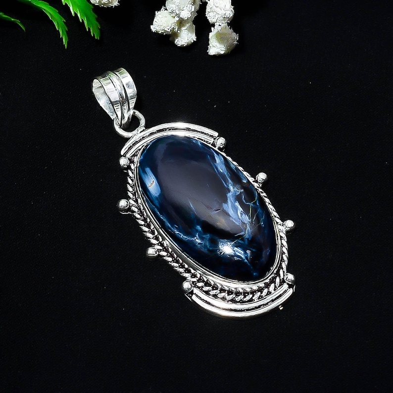 Puede incluir: Colgante ovalado con una piedra preciosa azul pulida, con vetas blancas, engastada en un marco de metal plateado. El marco tiene un borde decorativo en forma de cuerda y una argolla para una cadena. El colgante mide unos 5 cm de largo.