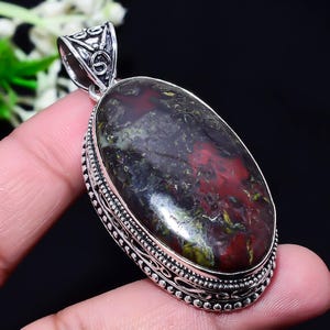 May include: Oval pendant with a large, polished gemstone. The stone features a mix of dark red, green, and black colors. The pendant is set in a decorative silver-colored metal frame with an ornate bail.